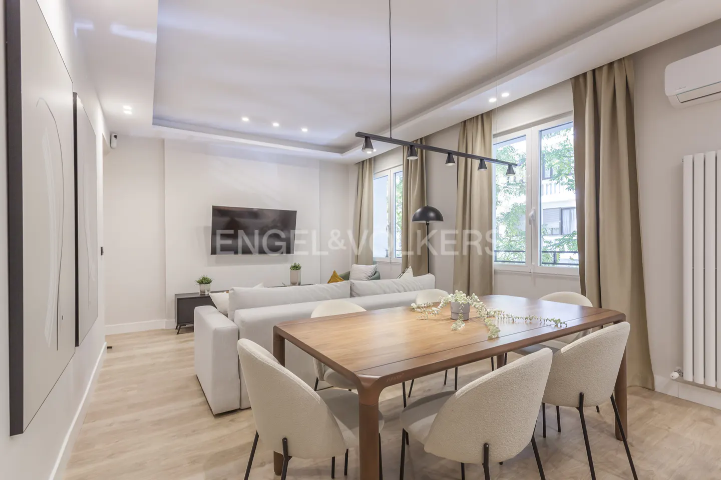 Bright, modern living and dining area with a wood table, white chairs, and a white sofa. Large windows with beige curtains.