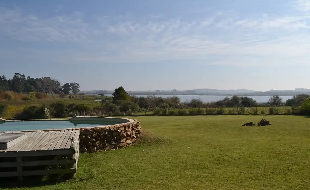 Outdoor pool with stone surround and wooden deck overlooks a green lawn and lake under a blue sky.