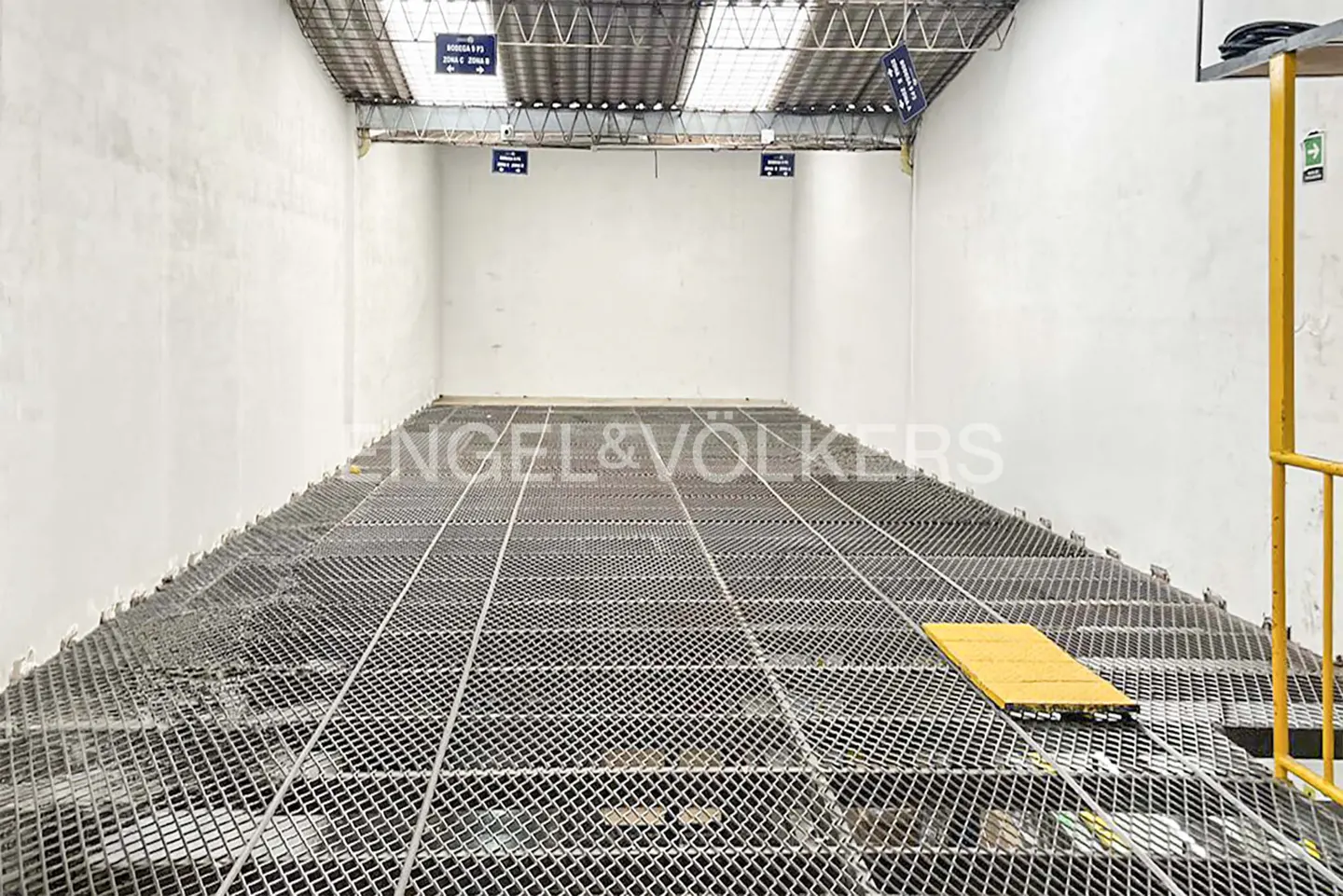 A warehouse interior with white walls and a metal grid floor. Yellow safety railing and signage are visible.