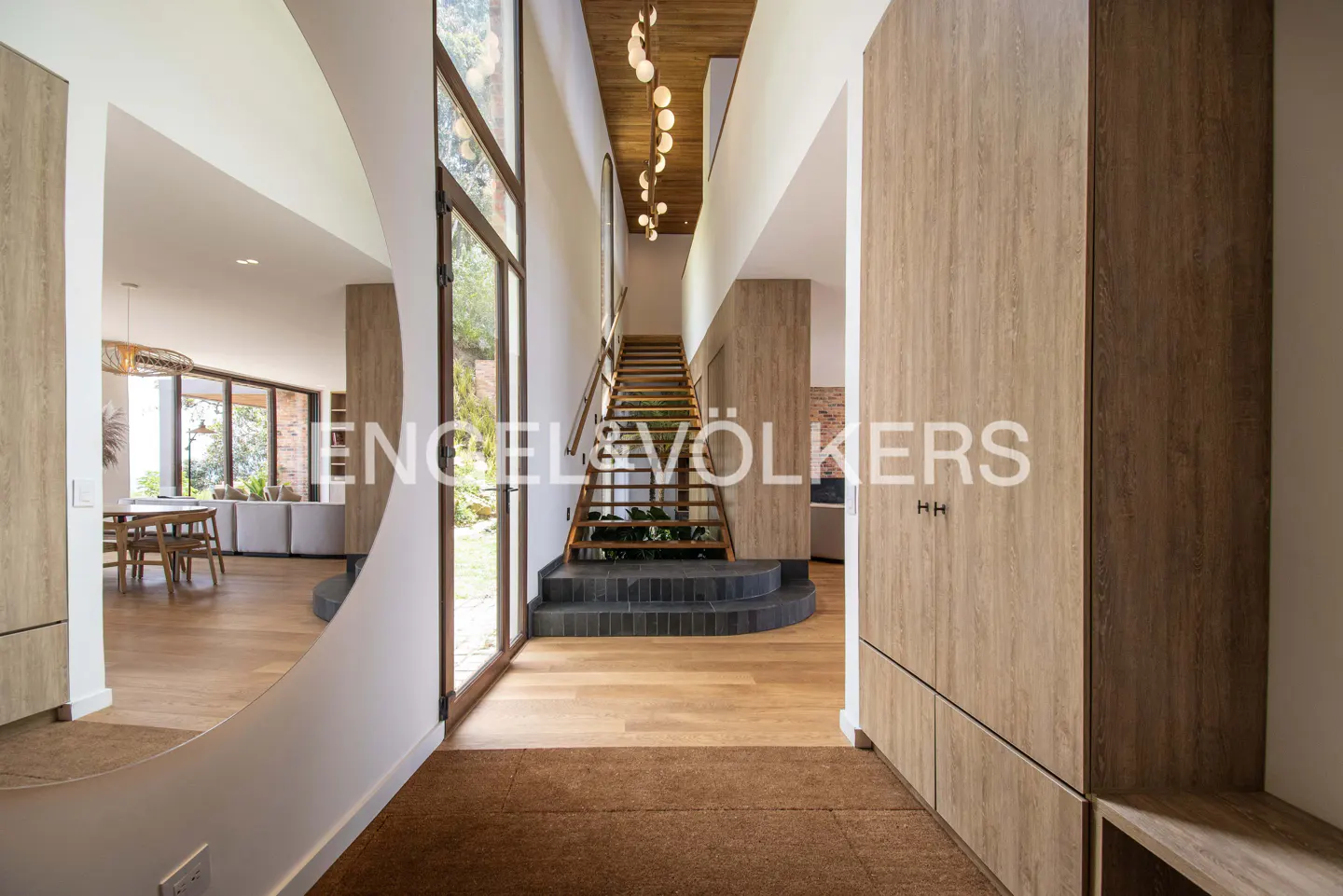 Entryway with wood floors, stairs, and a large round mirror reflecting a dining area. Tall windows let in natural light.