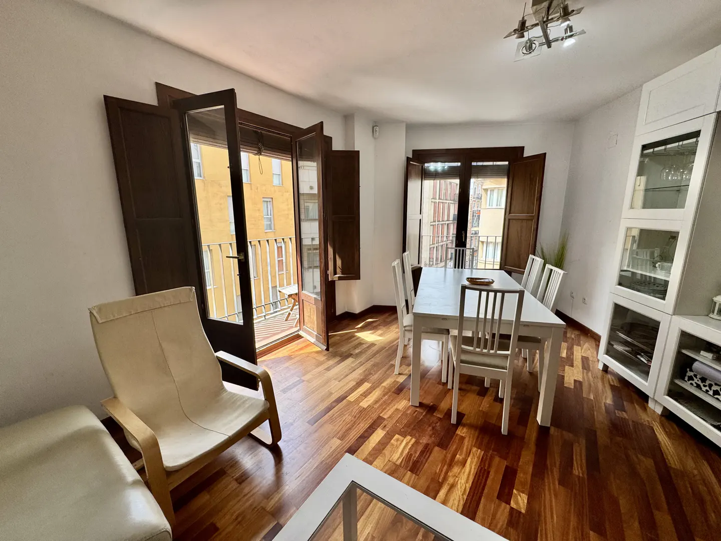 Bright living room with hardwood floors, white walls, and dark wood trim. Open balcony doors let in natural light. White dining table with chairs and a white cabinet.
