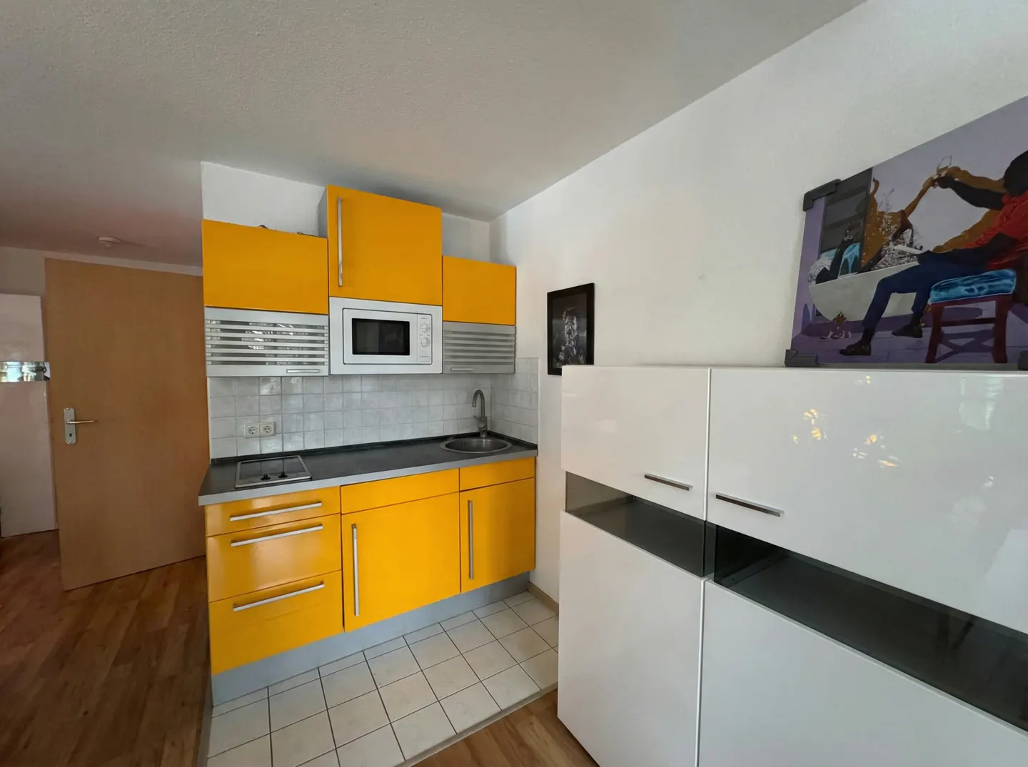 A compact kitchen with yellow cabinets, a microwave, and white storage units. A painting sits atop the storage.