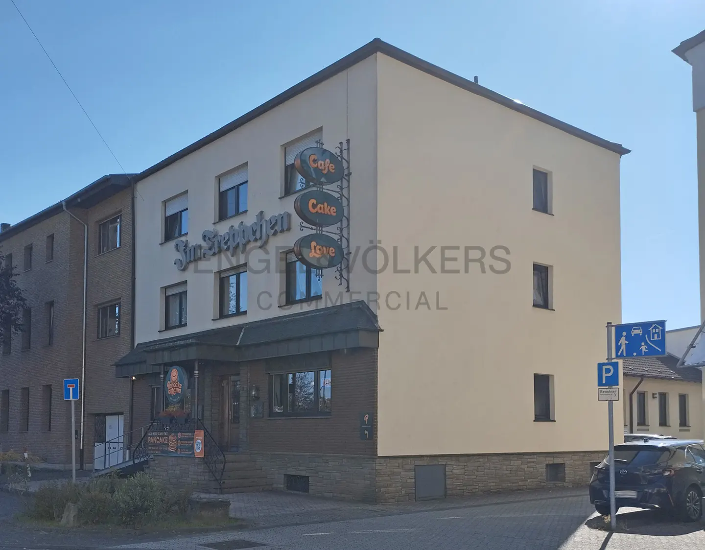 Exterior view of a two-story building with "Cafe Cake Love" sign and Engel & Volkers logo. A car is parked nearby.