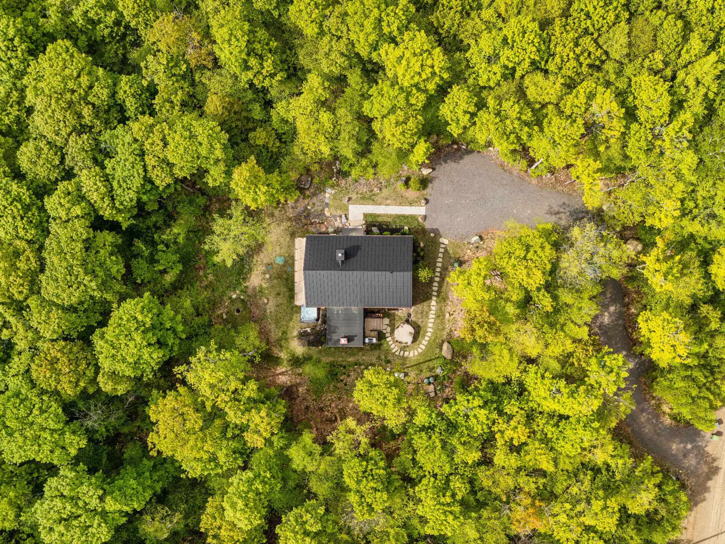 Aerial view of a house with a dark roof nestled in a dense, vibrant green forest with a gravel driveway and stone path.