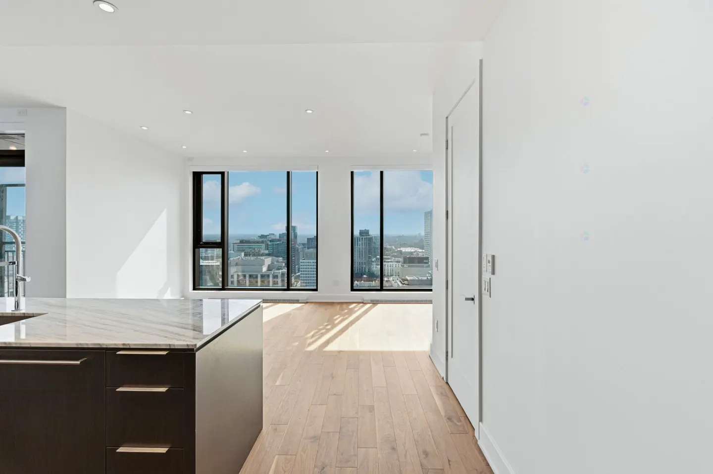 Bright, modern apartment interior with hardwood floors, white walls, and a kitchen island. Large windows offer a city view.