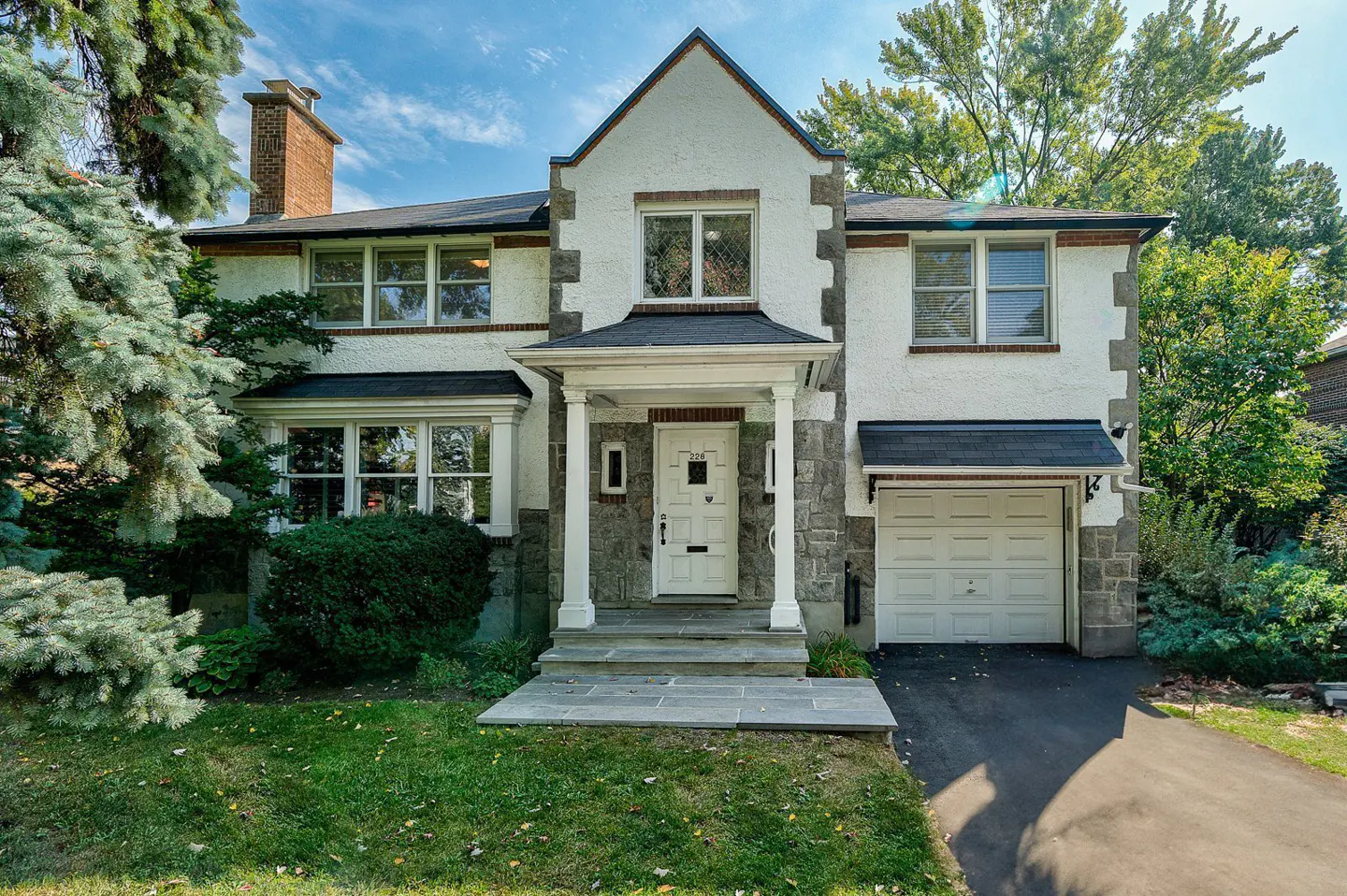 Two-story Tudor-style house with white stucco, stone accents, and a dark roof. Green lawn and trees surround the house.