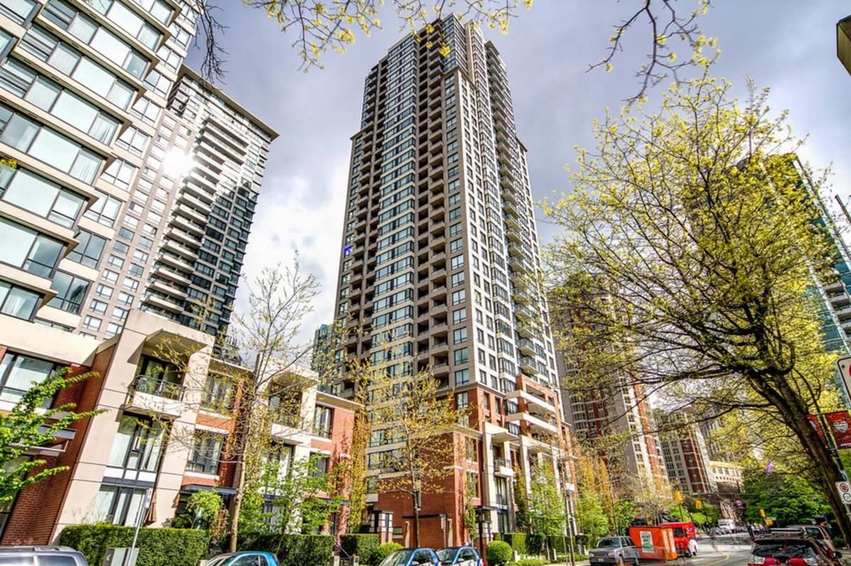 Exterior view of a tall condo building with balconies, surrounded by trees and other buildings in a city setting.