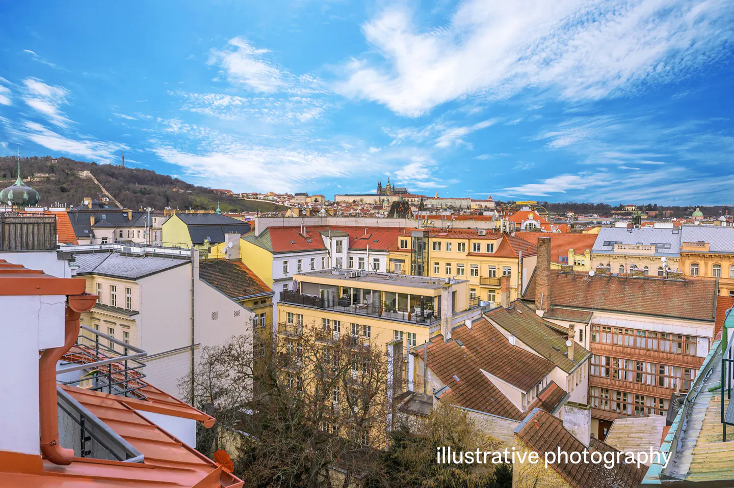 Cityscape view of Prague with red rooftops, yellow buildings, and Prague Castle in the background under a blue sky with white clouds.