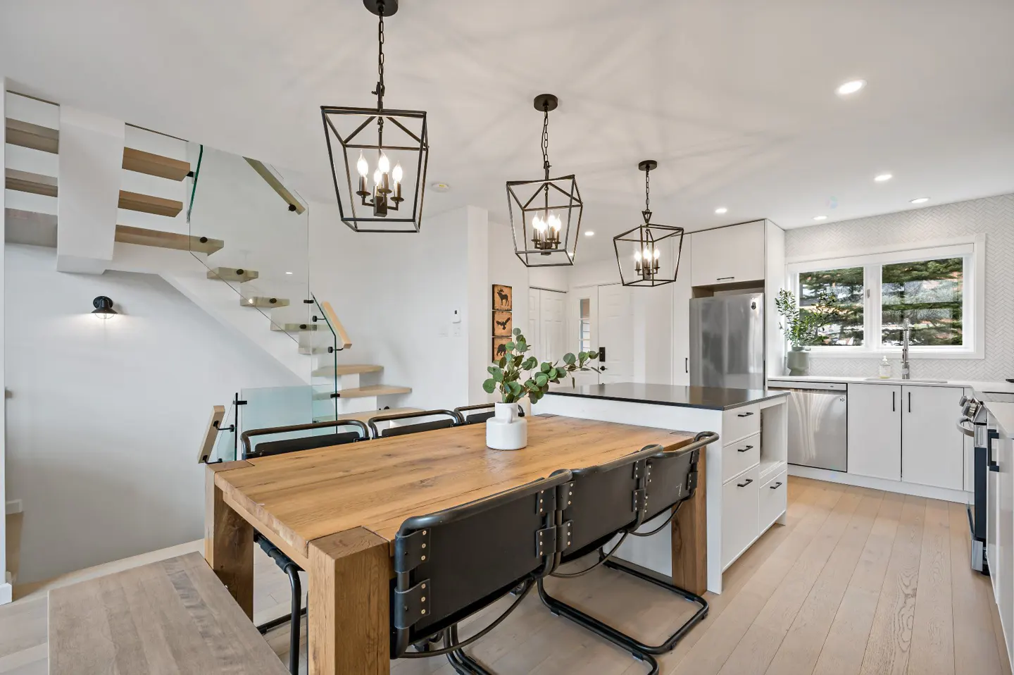 Bright, modern kitchen with a wood table, black chairs, and three black pendant lights. White cabinets and stainless steel appliances. Stairs with glass railing.