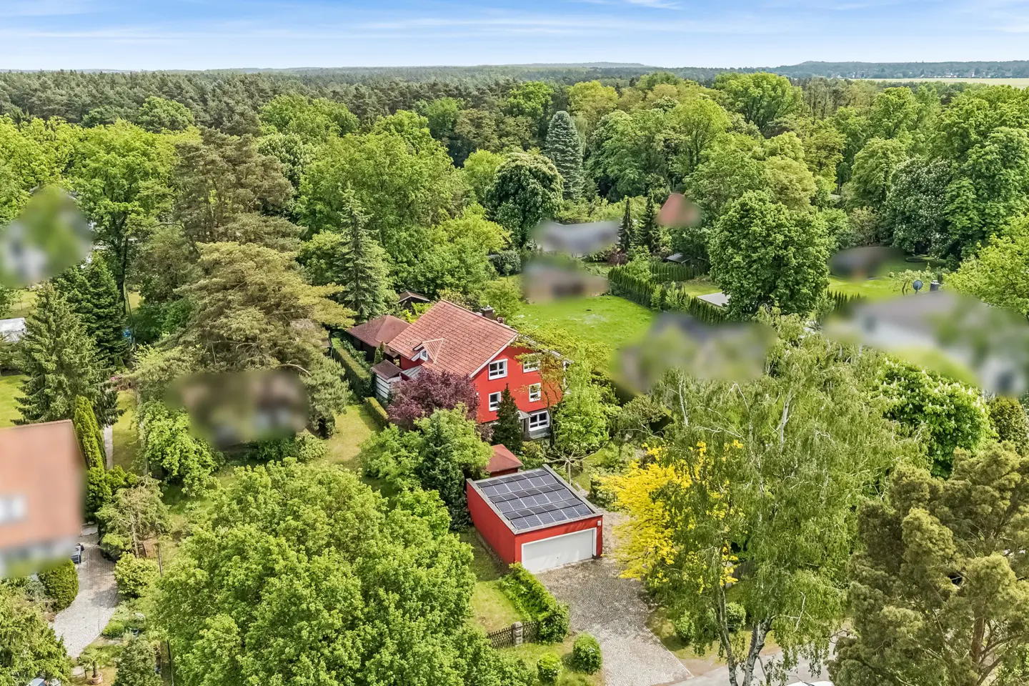 Aerial view of a red house with a tiled roof and a matching garage with solar panels, surrounded by lush green trees.