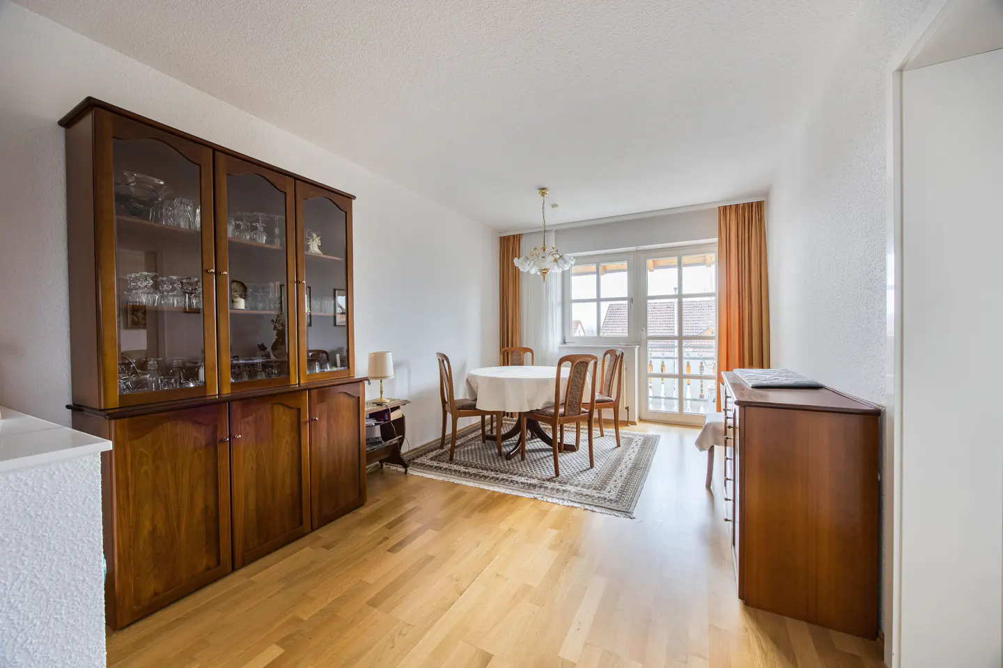 A dining room with light wood floors, a round table with four chairs, and a large wooden cabinet. Curtains frame a window.