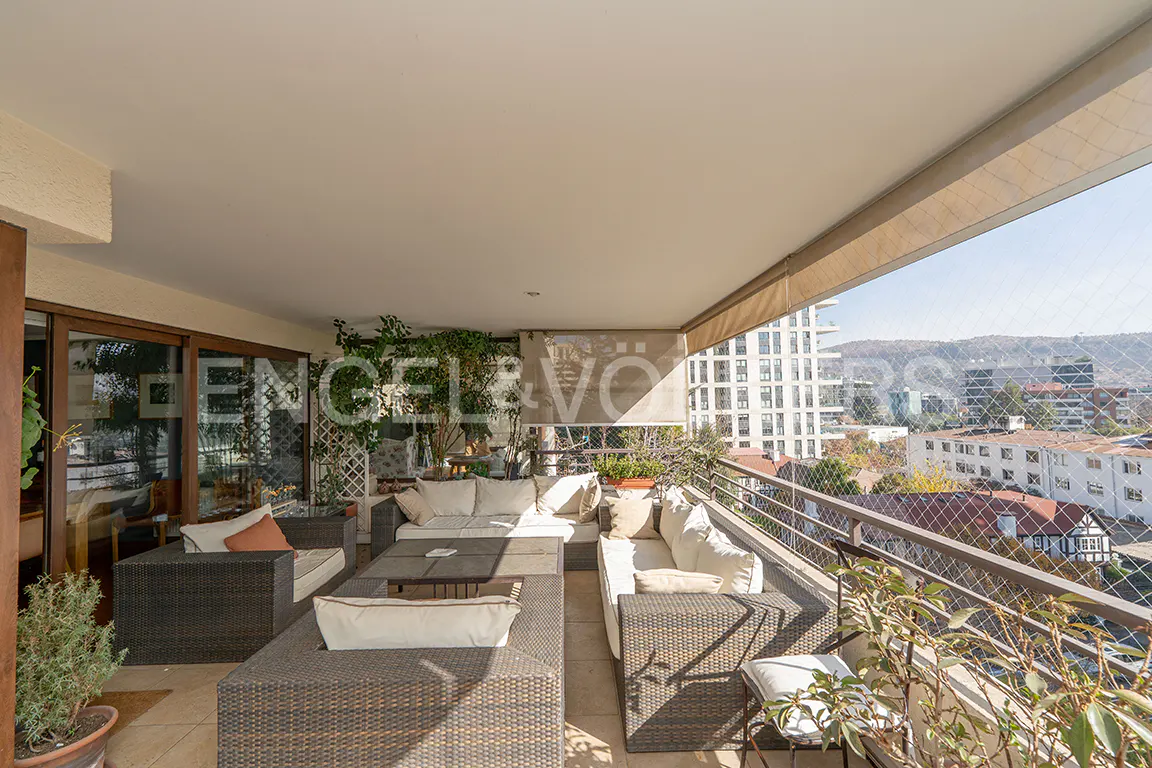 Spacious covered balcony with dark wicker lounge furniture, white cushions, and a city view with mountains in the distance.
