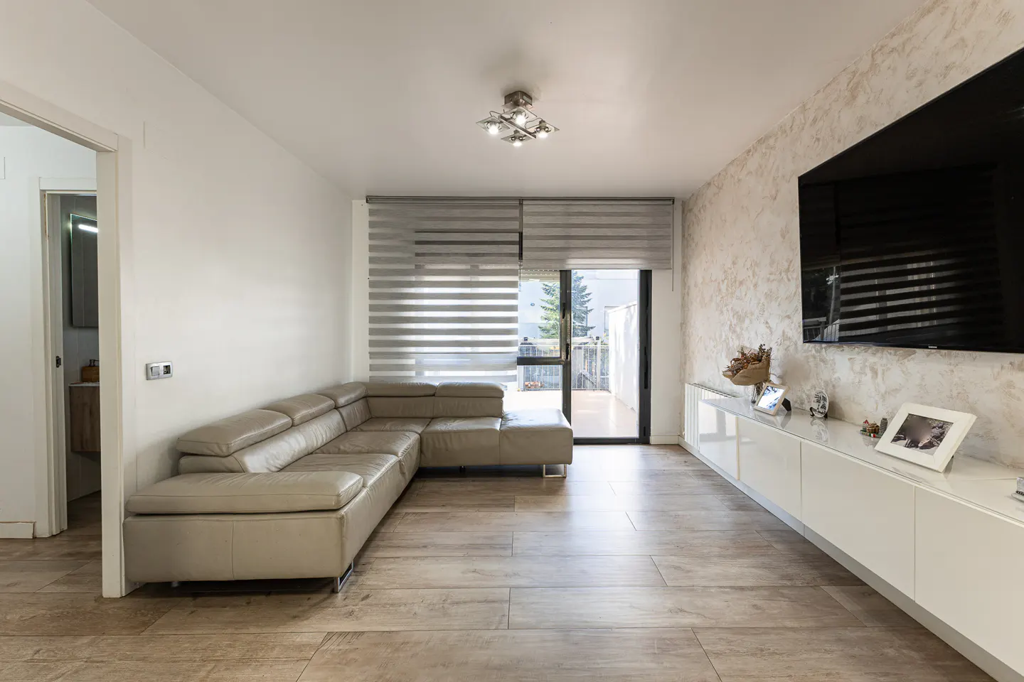 Living room with beige L-shaped sofa, wood-look tile floor, and white media console under a large TV. Blinds cover a sliding glass door.