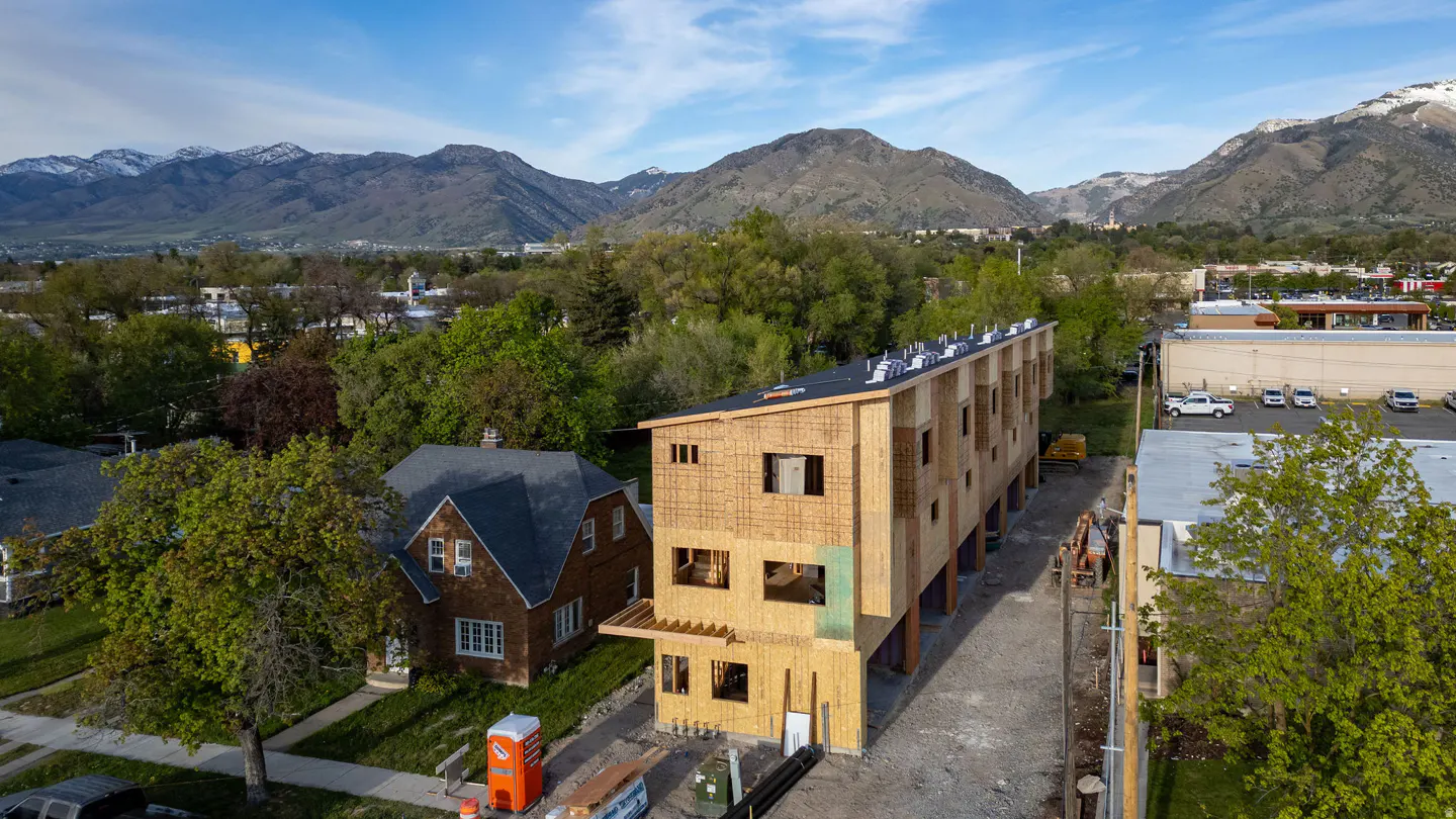 Aerial view of a new townhouse construction with exposed wood framing, set against a backdrop of green trees and snow-capped mountains.