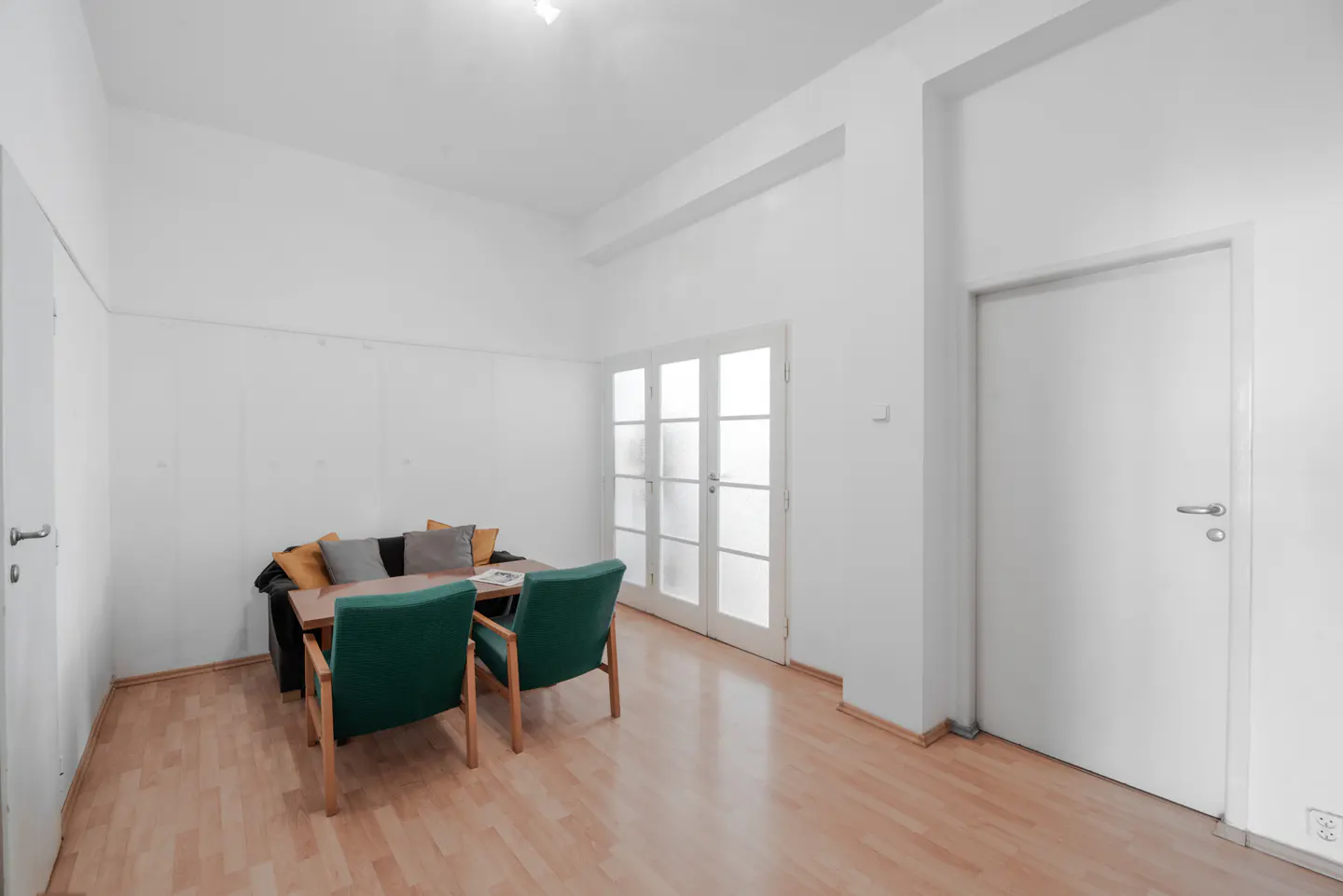 Bright room with light wood floors, a dark sofa with colorful pillows, two green armchairs, and glass-paned double doors.