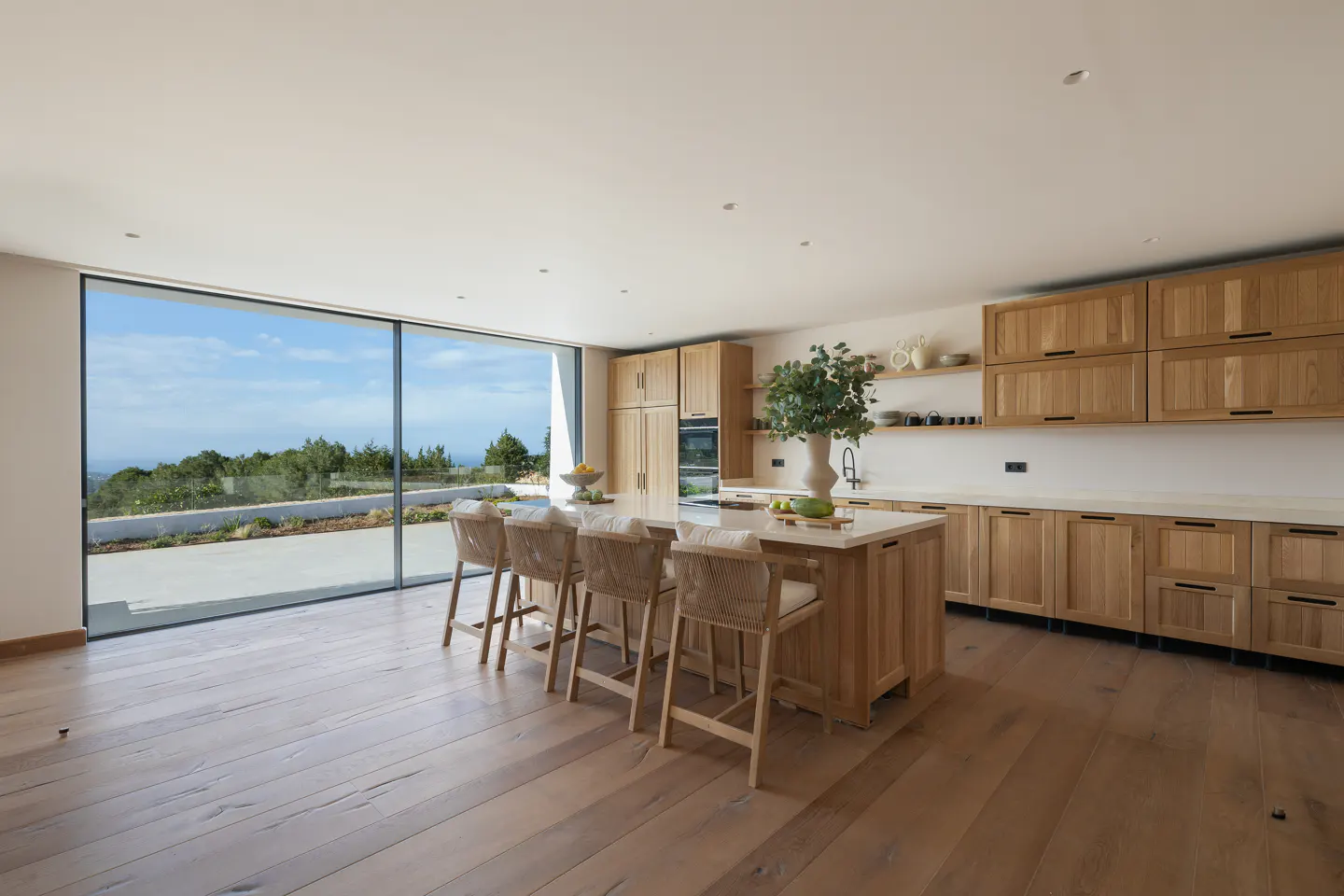A bright, modern kitchen with light wood cabinets, island with stools, and a large window with a view of the ocean.