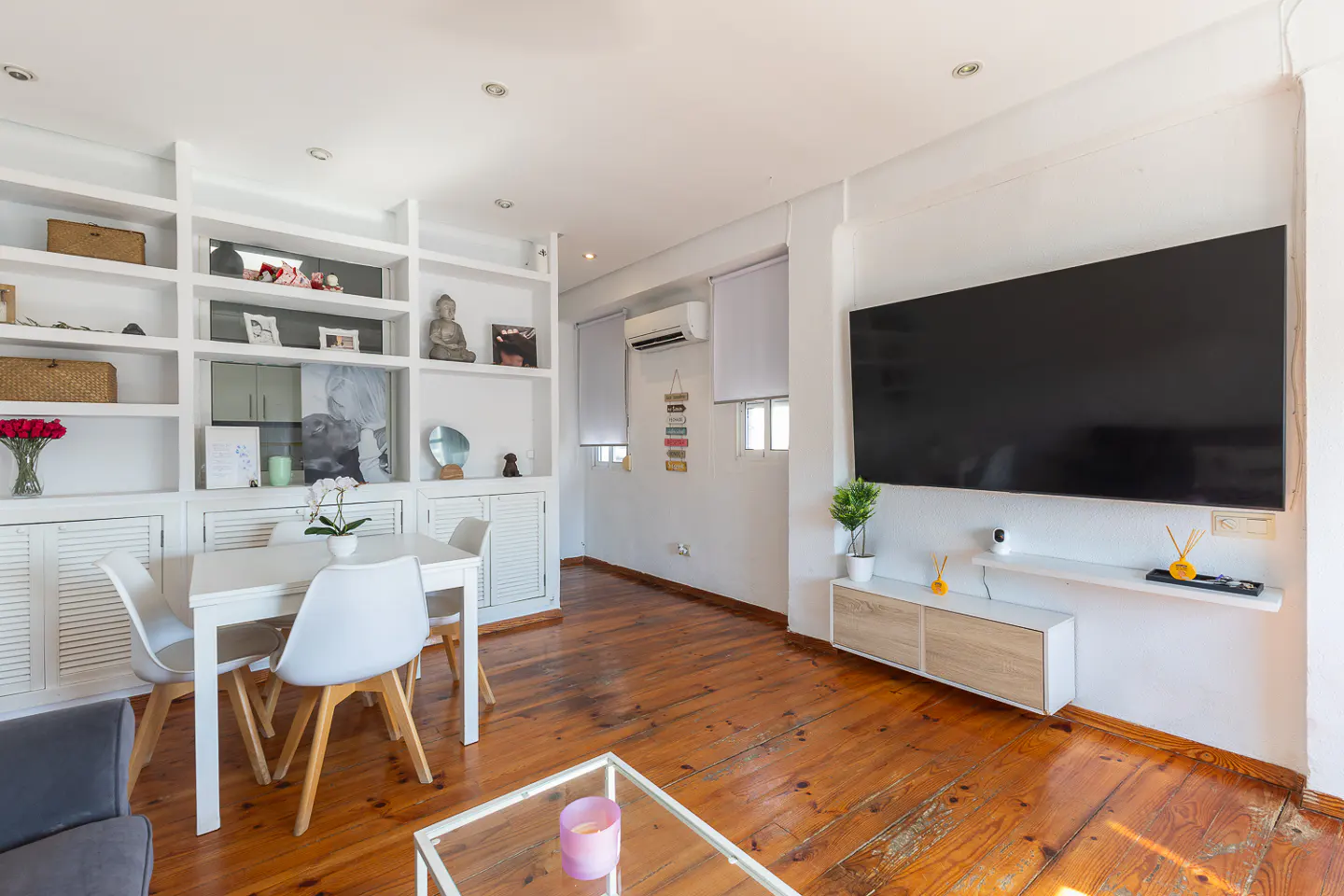 Bright living room with wood floors, white walls, and built-in shelves. A white table and chairs sit near the shelves, and a large TV hangs on the opposite wall.