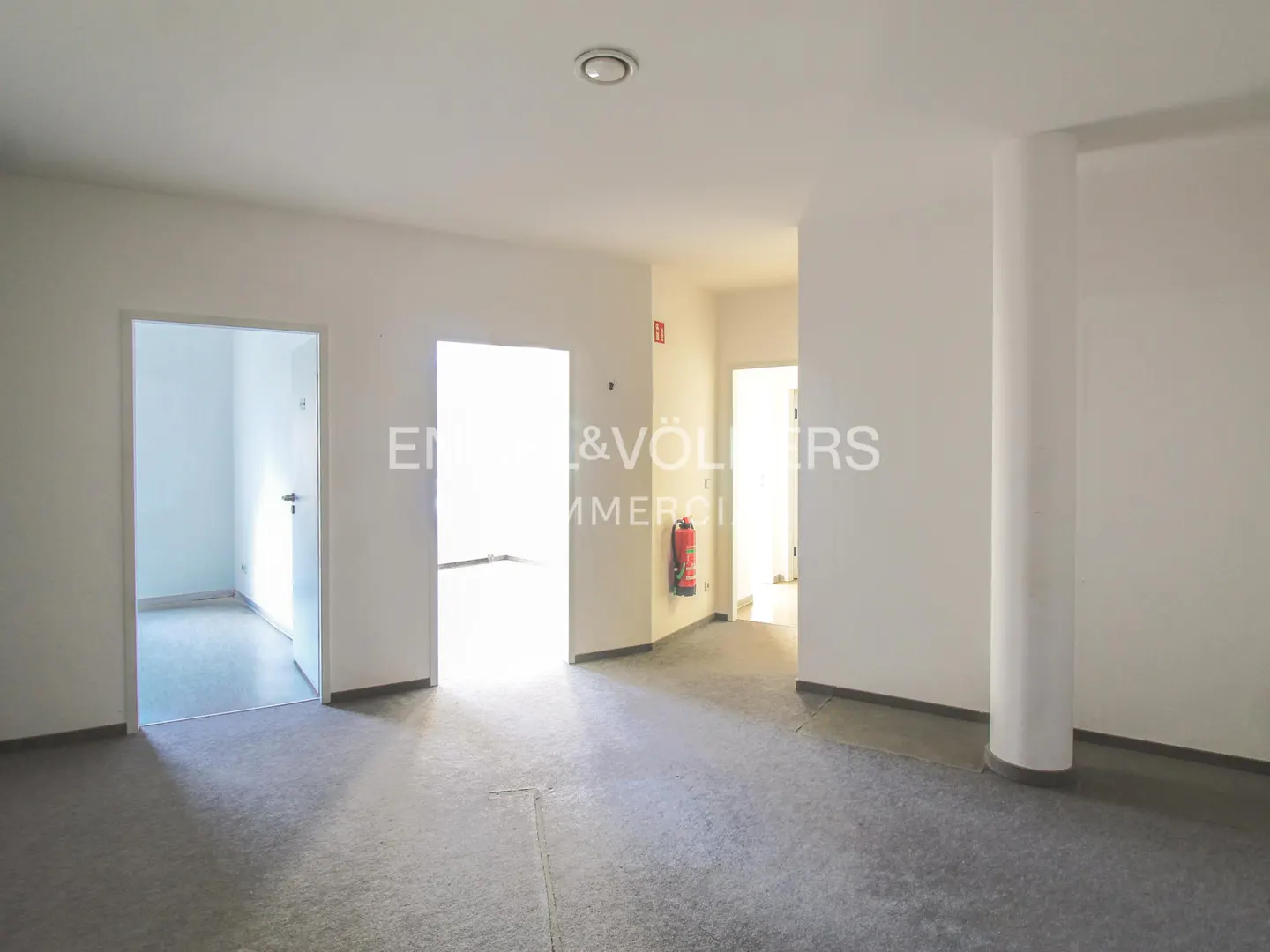 Empty room with gray floor, white walls, and multiple doorways. A red fire extinguisher is visible.