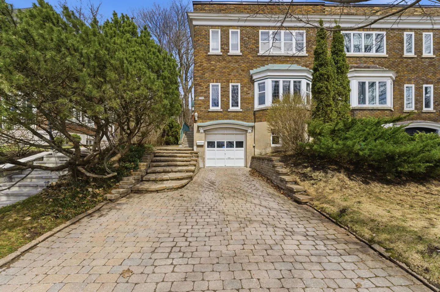 Three-story brick townhouse with a white garage door and a stone driveway. Green trees and shrubs surround the house.