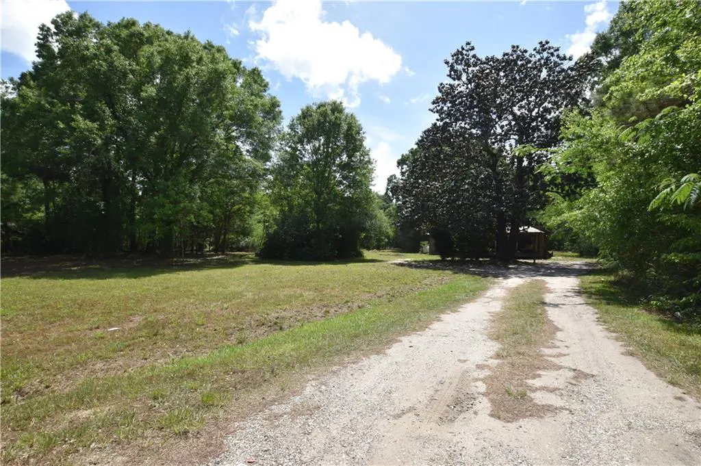 Gravel driveway leading to a wooded lot with a small wooden structure in the background. Green grass and trees surround the property.