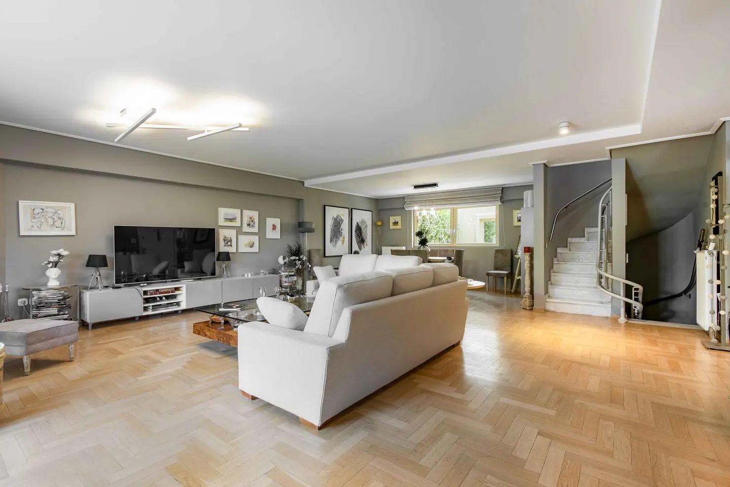 A spacious living room with a white sofa, TV, and a staircase. The walls are gray, and the floor is parquet.