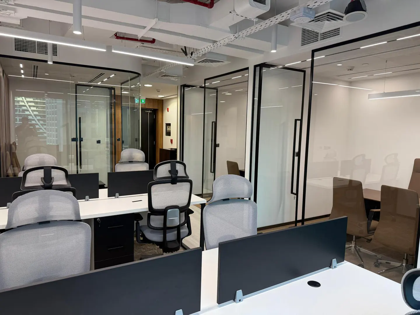 Bright office space with white desks, gray chairs, and black dividers. Glass-walled conference rooms are visible in the background.