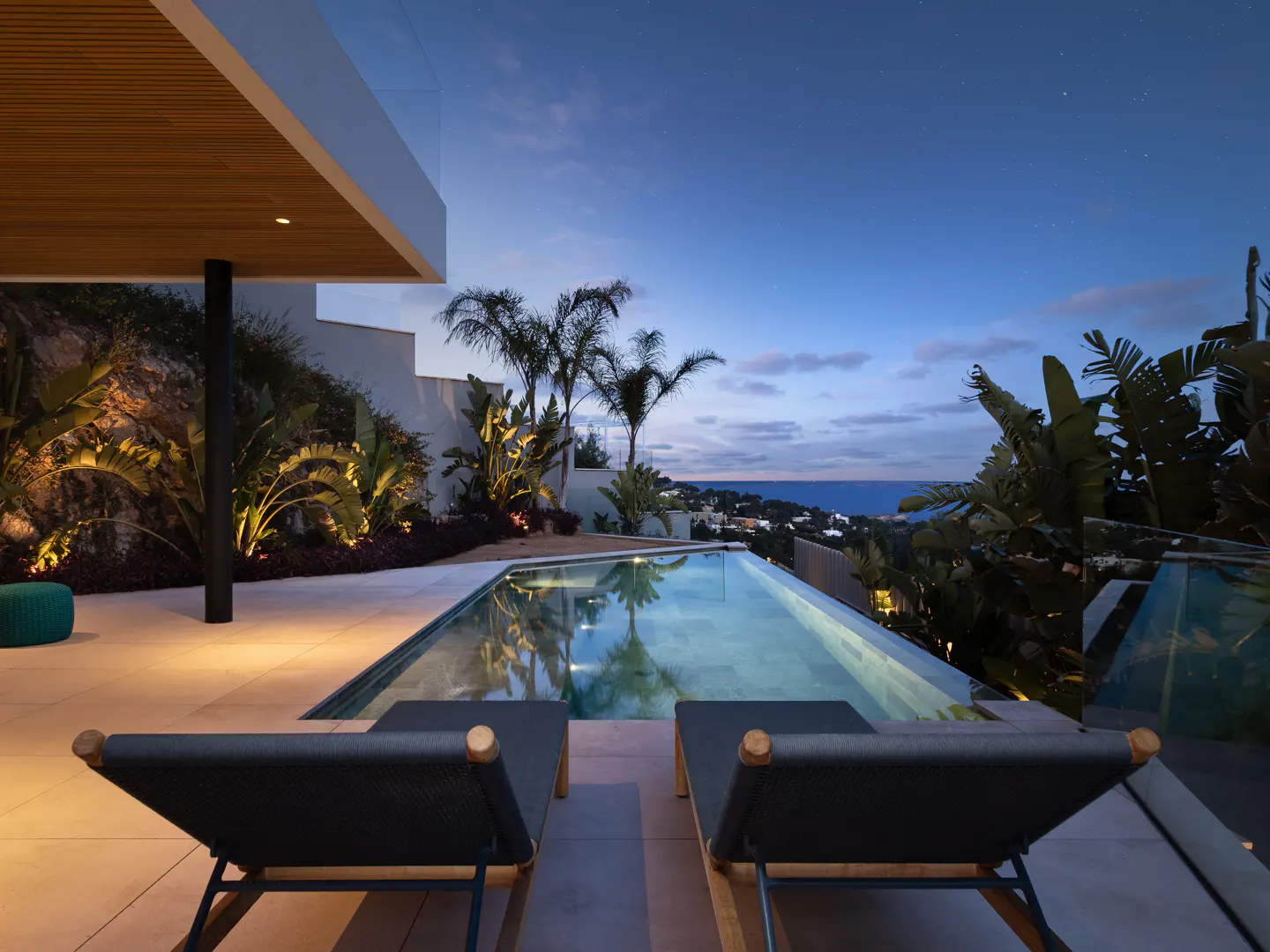 Outdoor pool area at dusk with two lounge chairs in the foreground, palm trees, and ocean view.
