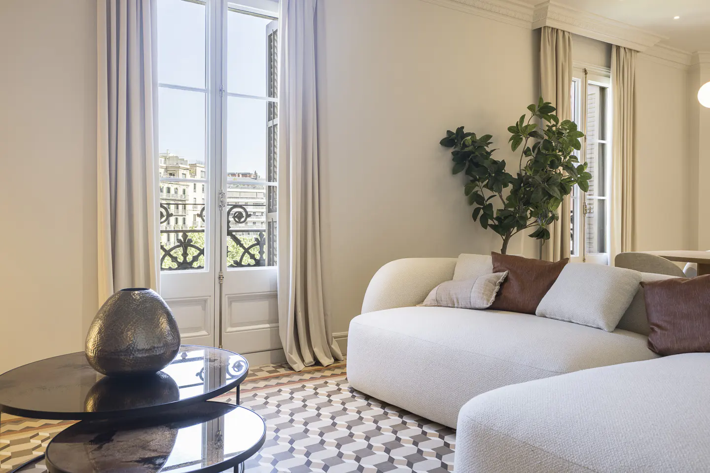 Bright living room with a white sofa, brown pillows, and a plant. A window with curtains overlooks a city. Two black tables sit on a patterned floor.