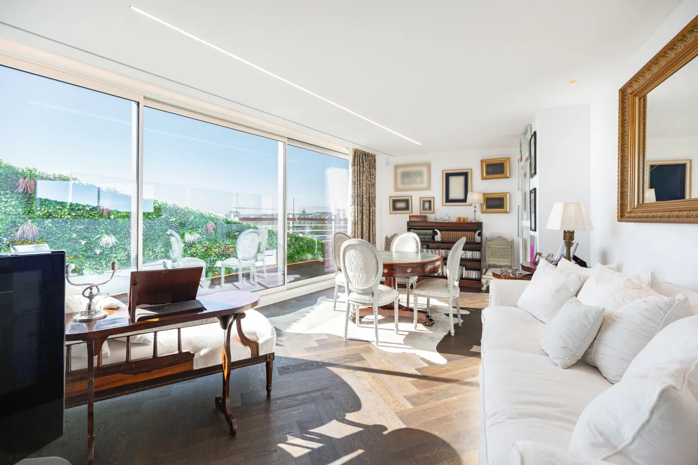 Bright living room with a white sofa, a wooden desk with a laptop, and a dining table with white chairs. Large windows overlook a green terrace.
