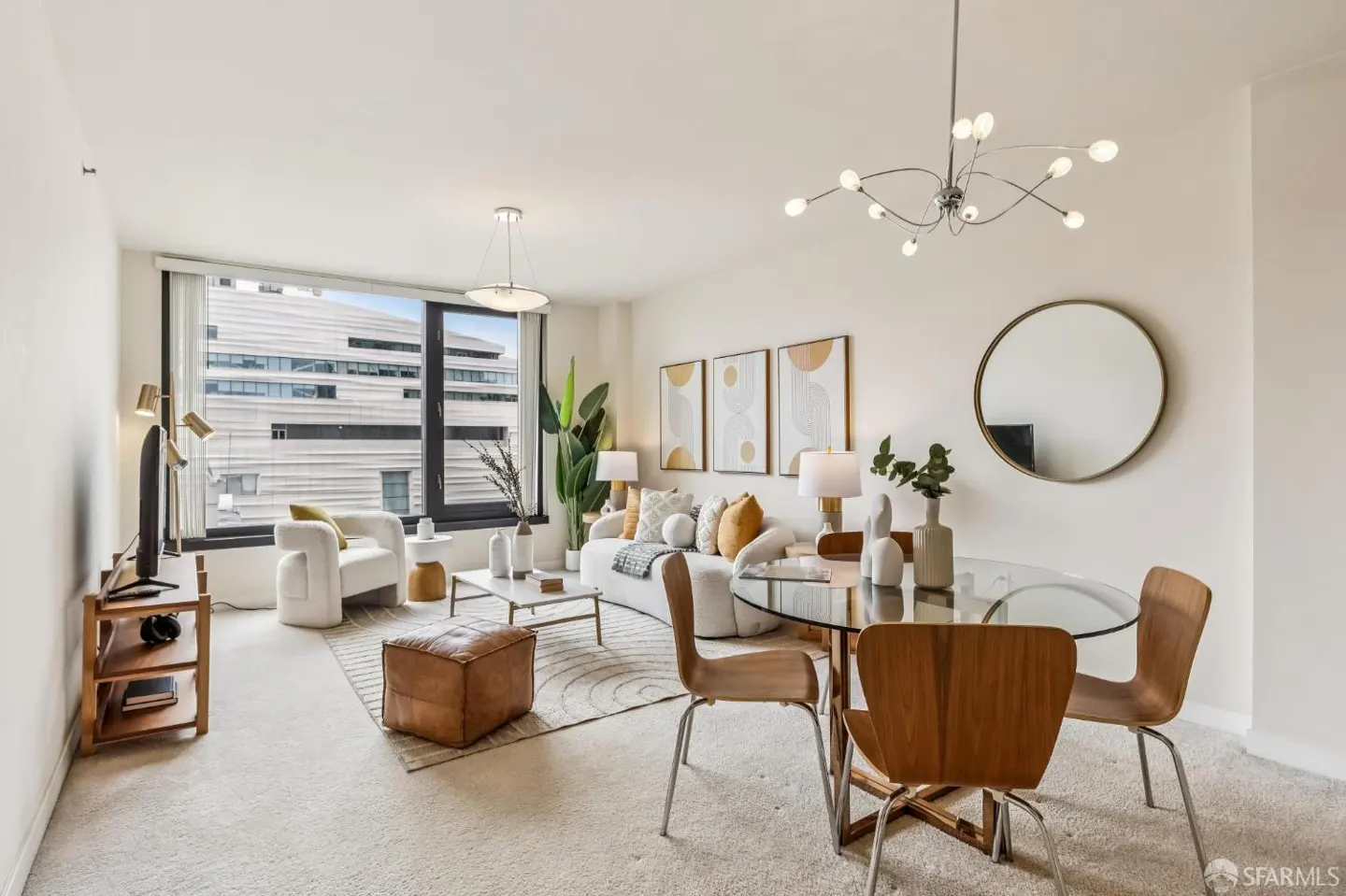 Modern living room with a white sofa, leather ottoman, glass dining table, and large window overlooking a city building.