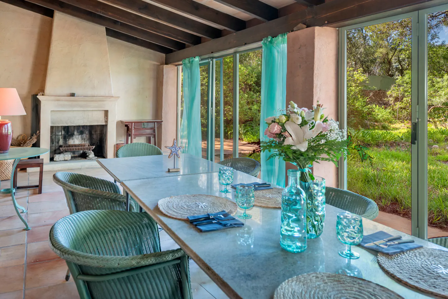 Dining room with a long table set for a meal, wicker chairs, and a view of a green garden through glass doors.