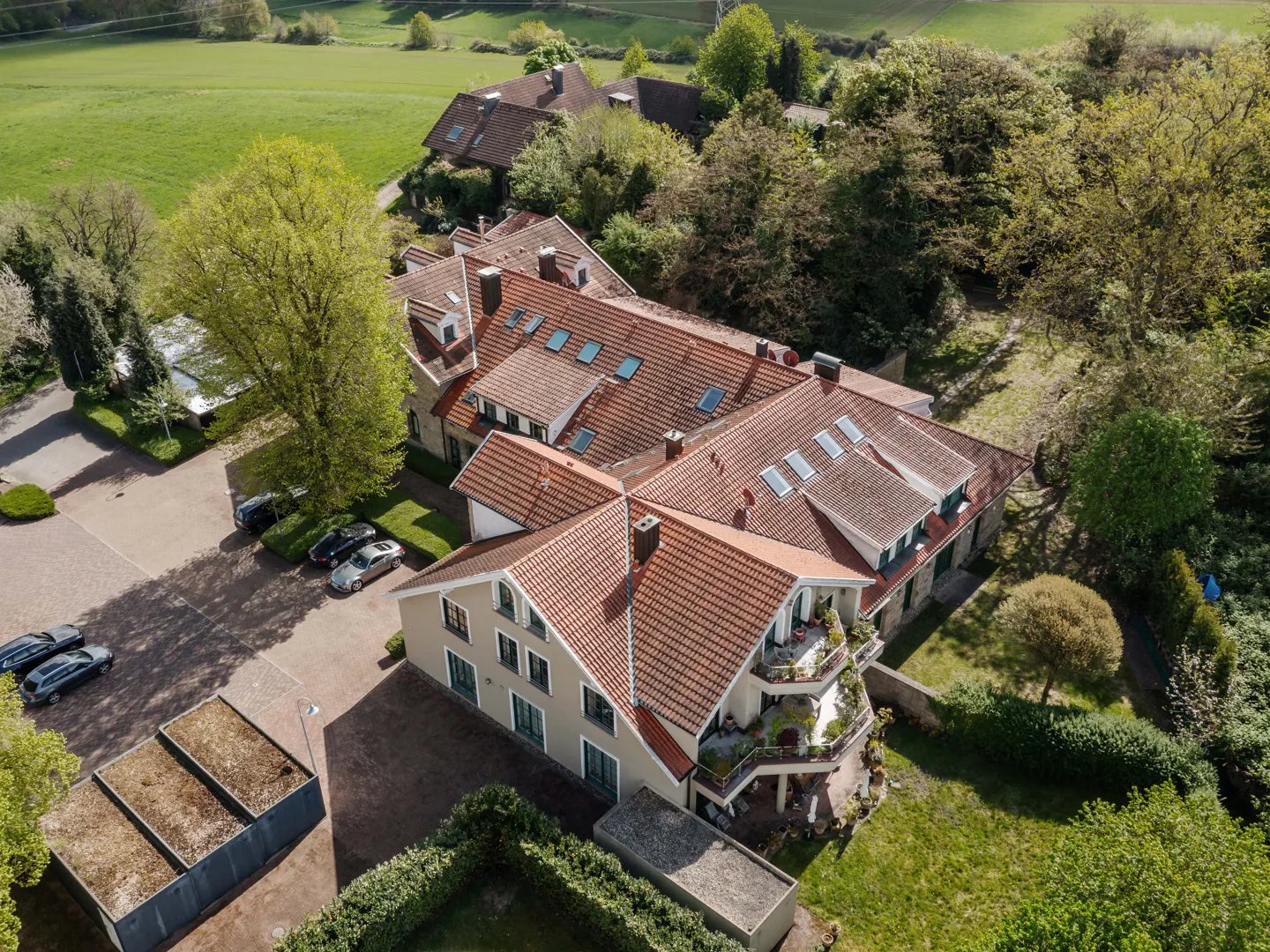 Aerial view of a large, beige building with a red tile roof, surrounded by green trees and grass. Cars are parked nearby.