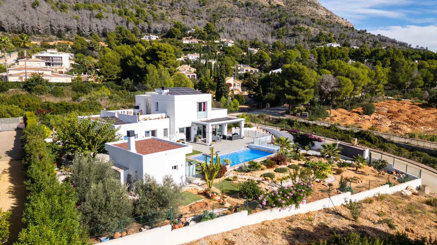 Aerial view of a modern white house with a pool, surrounded by lush greenery and a mountain backdrop.