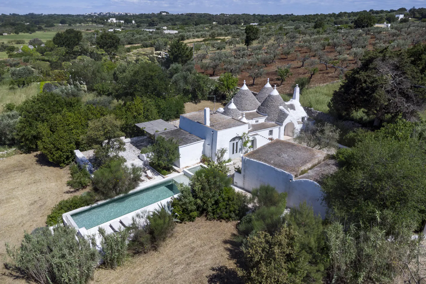 Aerial view of a white Trulli house with conical roofs, a pool, and olive trees in the background.