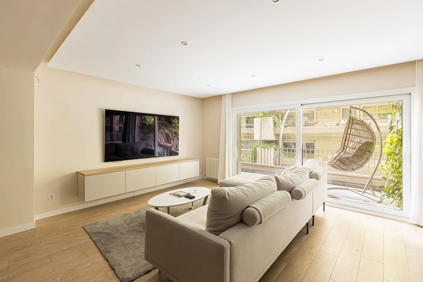 Living room with a beige sofa, gray rug, and TV above a white cabinet. A hanging chair is visible through the sliding glass doors.