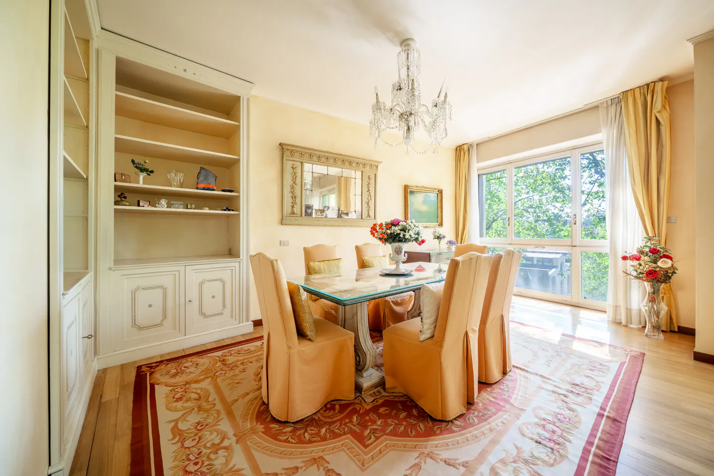 A dining room with a glass table, peach chairs, a chandelier, and a large window.