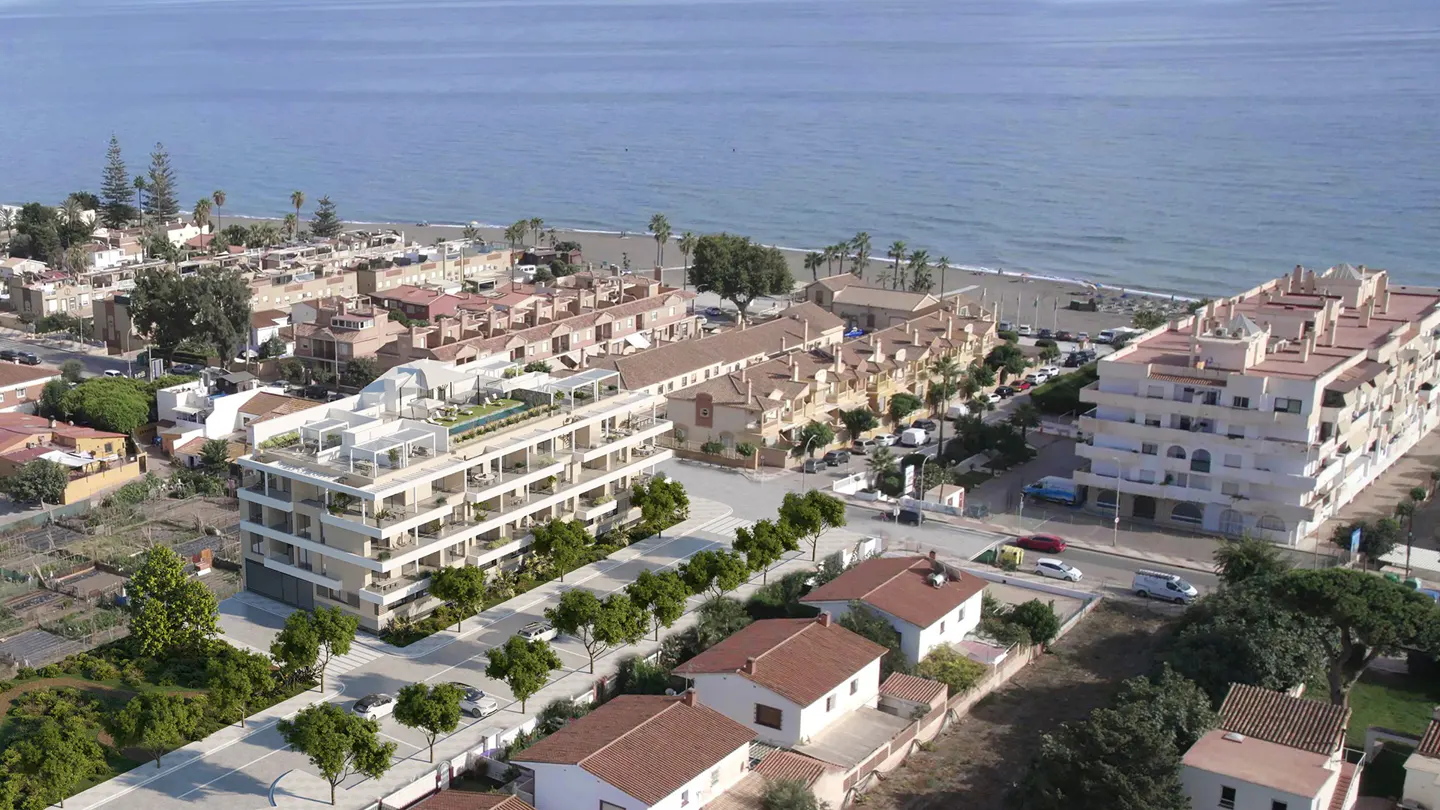 Aerial view of a modern, multi-story apartment building with a rooftop pool, near a beach and other residential buildings.