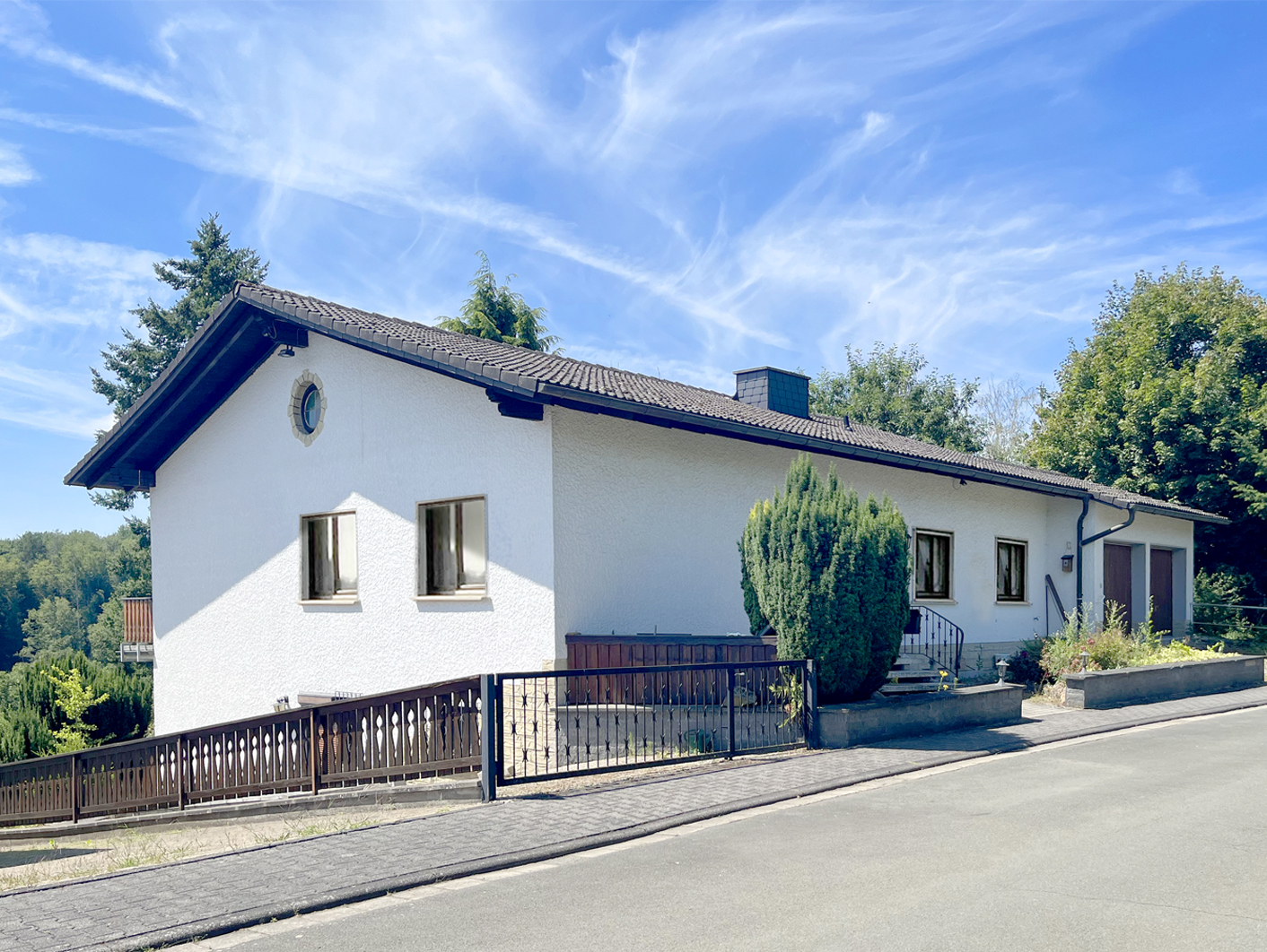 White single-story house with a dark tiled roof, a fenced yard, and a garage, set against a bright blue sky.