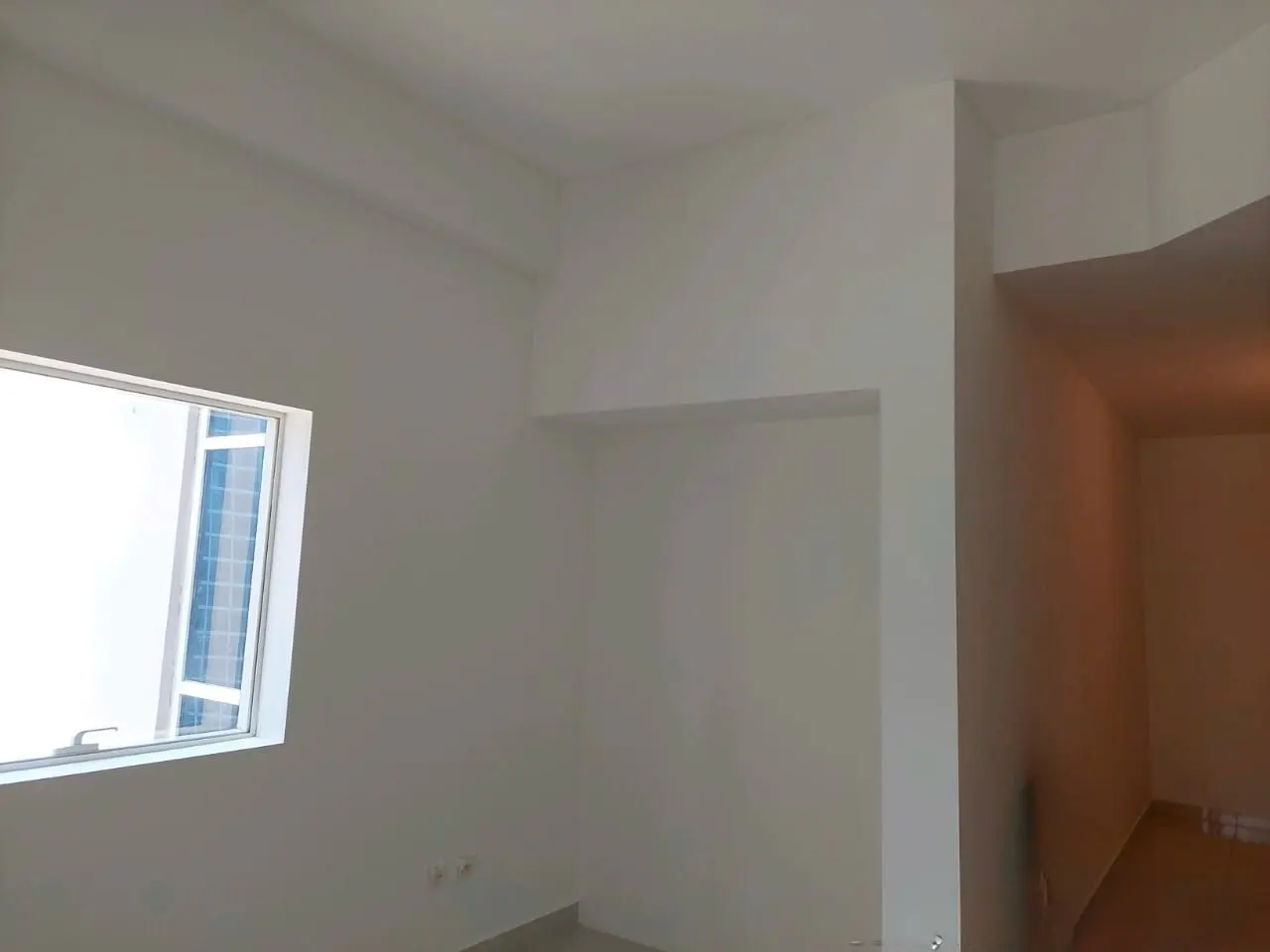 Bright, empty room with white walls, a window showing a blue building, and an open doorway to another room.