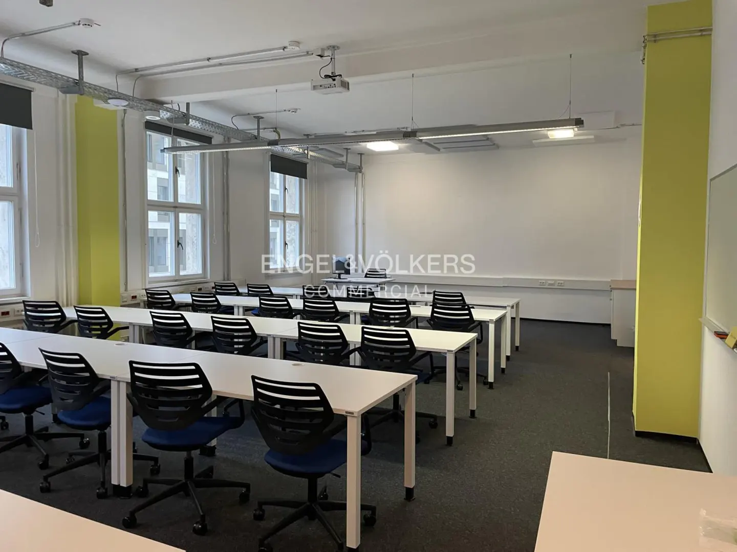 Classroom view with rows of white tables and blue chairs, projector on ceiling, and yellow accent wall.