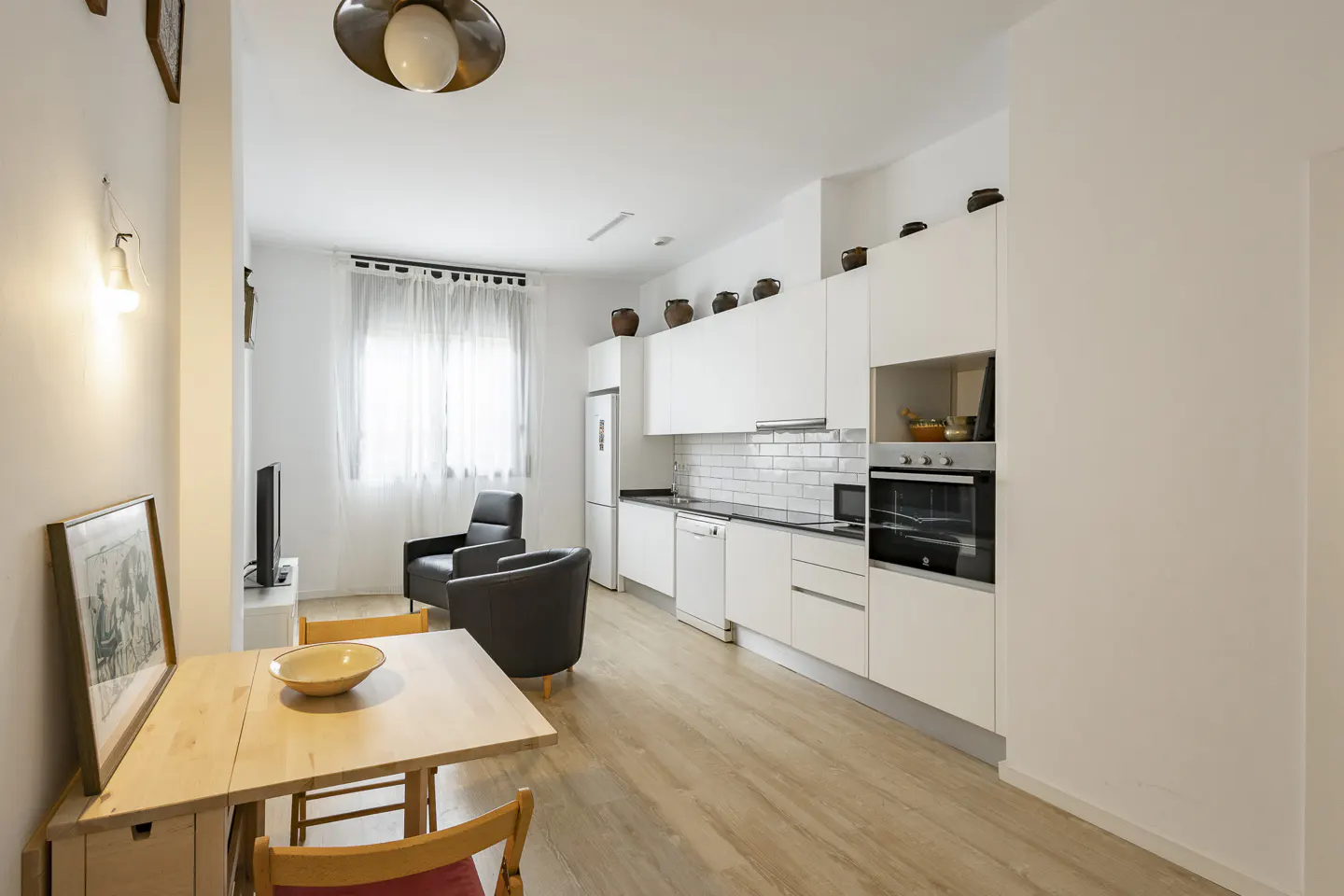 Modern open-plan kitchen and living area with white cabinets, light wood floors, a dining table, and two black armchairs.