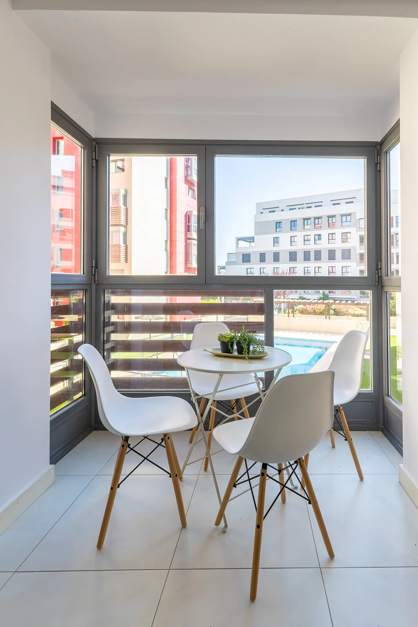 Balcony with white table and chairs. Outside view of buildings and pool through gray framed windows. Plants on table.