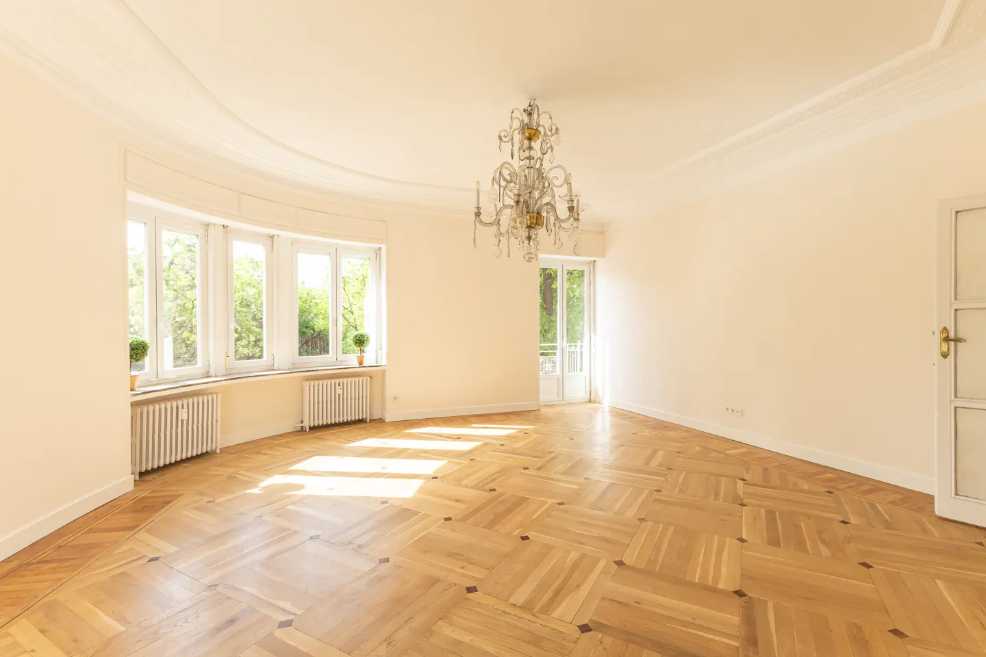 Bright, empty room with parquet floors, white walls, and a crystal chandelier. Sunlight streams through a bay window overlooking greenery.