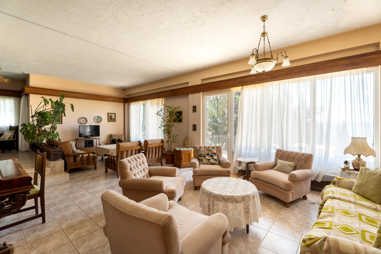 A spacious living room with beige armchairs, tile flooring, and large windows with sheer white curtains. A chandelier hangs from the ceiling.
