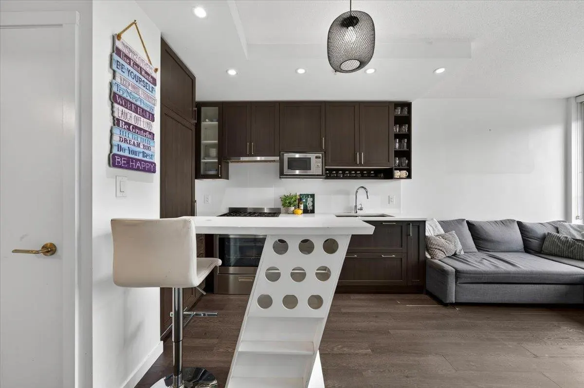 A modern kitchen with dark wood cabinets, white countertops, and a white island with a bar stool. A gray sofa is visible in the background.