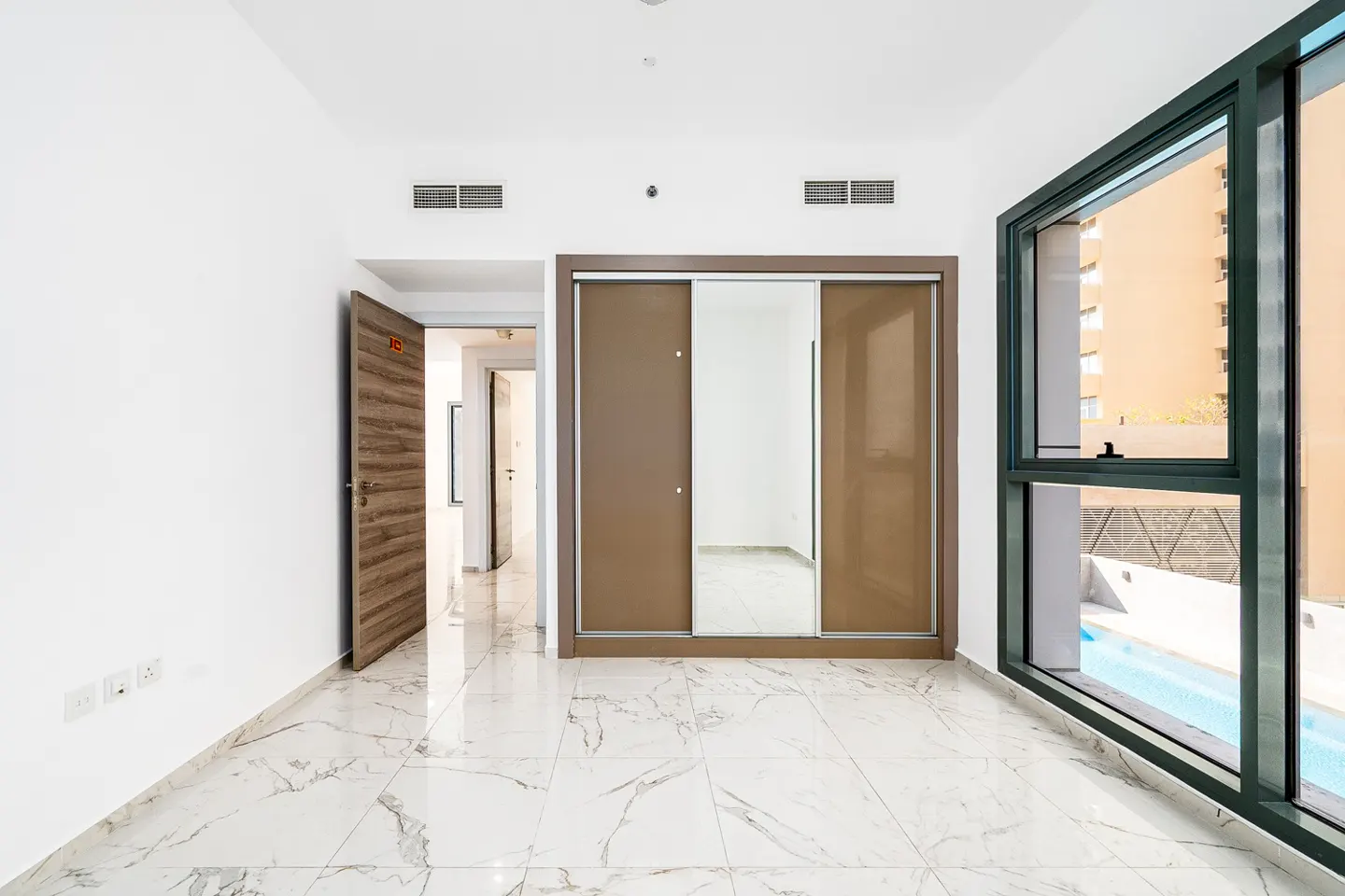Bright, empty room with white walls, marble floors, and a large window overlooking a pool. A brown sliding door closet and an open wooden door are visible.