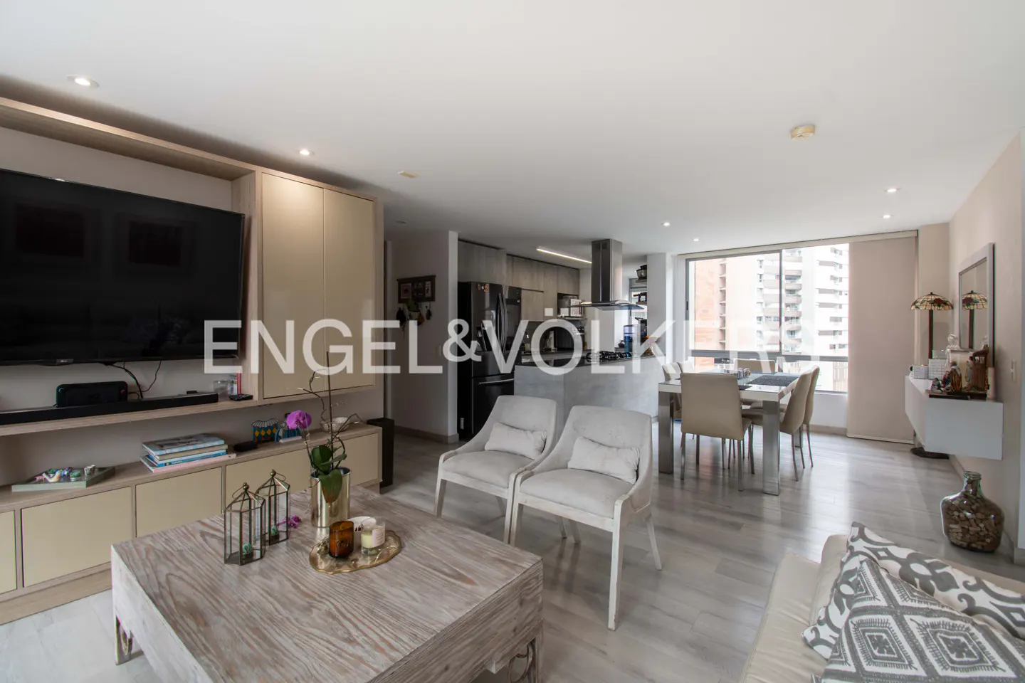 Bright, open-concept living space with a TV, kitchen, dining area, and large windows. Neutral colors and modern furniture. Engel & Völkers logo.