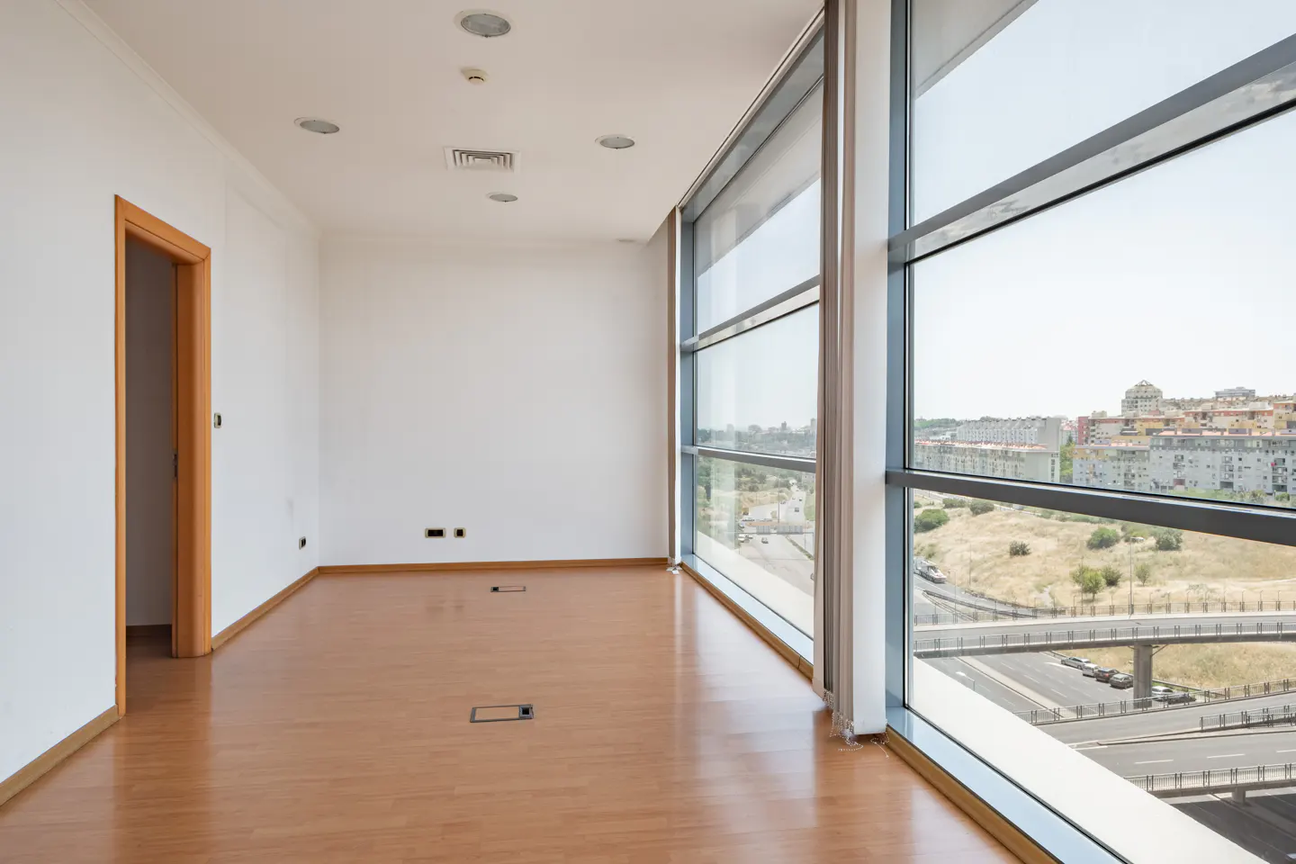 Empty office space with white walls, wood floor, and large windows overlooking a city and highway.