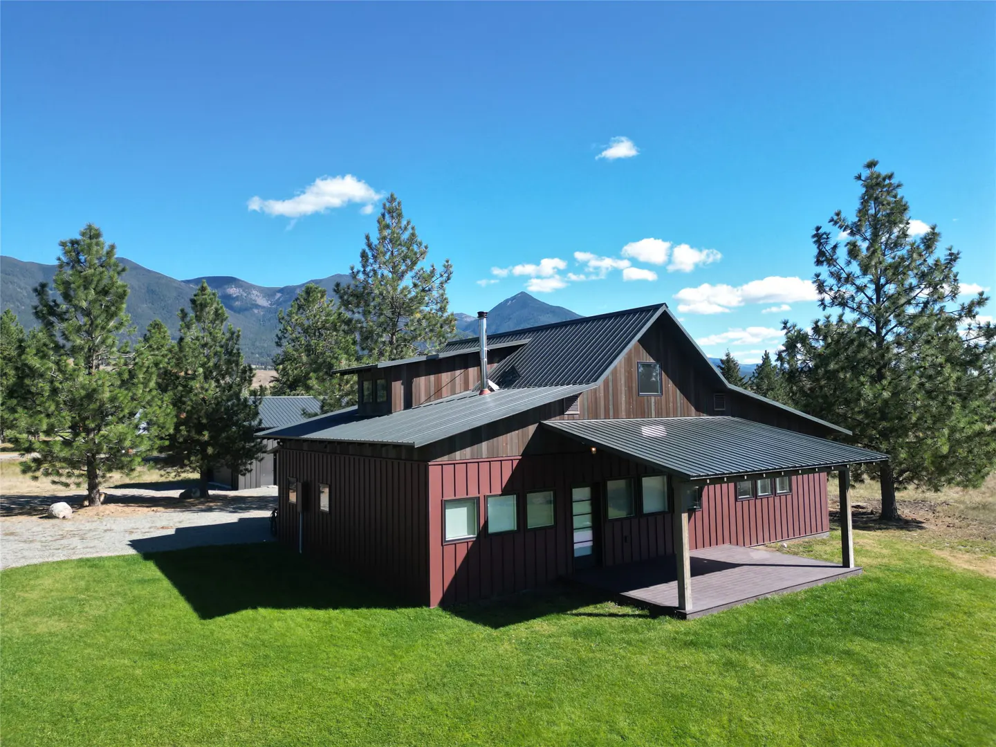 Exterior view of a barn-style house with a metal roof, red siding, and a covered porch, set against a backdrop of mountains and a blue sky.