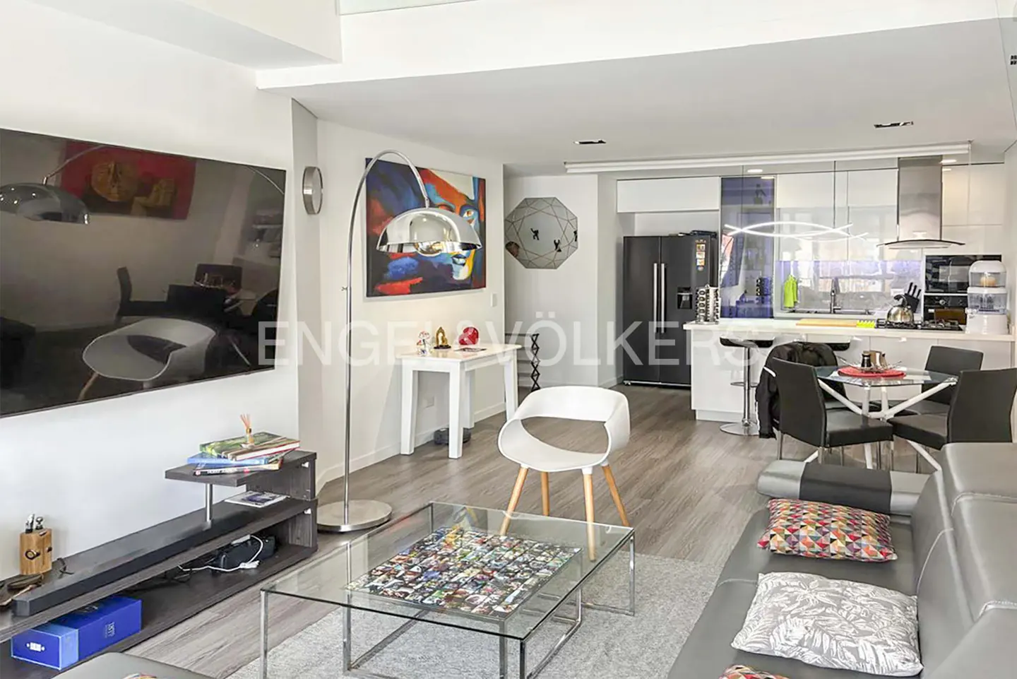Open-concept living space with gray sofa, glass coffee table, and modern kitchen with black fridge. Artwork and a floor lamp add style.
